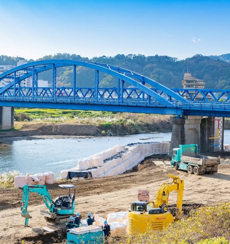 河川敷で重機が工事を行う様子。川には青い橋が架かっている。